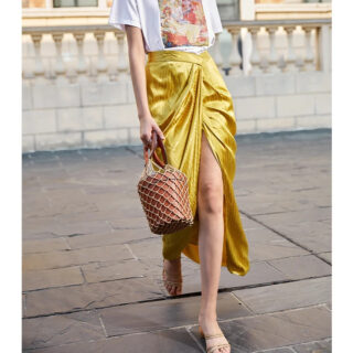 Photo d'une femme qui marche un panier rond à la main et qui porte une jupe drapée jaune fendue devant, un tee-shirt blanc et des sandales
