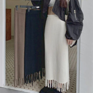 Photo d'une femme debout portant une jupe droite longue en tricot beige à franges un top blanc et un blouson court noir et tenant deux cintres avec les mêmes jupes en noire et en marron