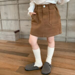 On voit le bas du corps d'une petite fille qui porte une jupe en coton épais marron, avec des chaussettes hautes blanches et des chaussons gris.
