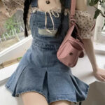 Photo d'une jeune femme assise sur un rebord d'une fenêtre et portant une jupe salopette en jean bleu plissée avec poche déchirée devant et un petit sac rose en bandoulière