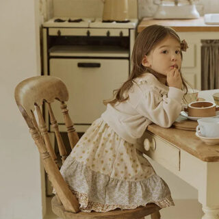 On voit une petite fille brune assise à la table d'une cuisine rétro dans les tons beiges. Elle porte une jupe à fleurs rétro.