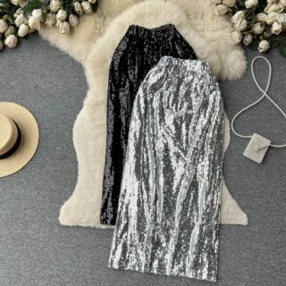 Photo de deux jupes à sequins taille haute élastique, une argente sur une noir, sur un tapis en poils blanc à côté d'un chapeau de paille et d'un sac à main.