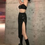 Photo d'une jeune femme debout les bras en l'air et portant une jupe fendue noire longue fermée par de grosses épingles de sûreté, un crop top noir et des bottes noires