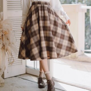 On voit le bas du corps d'une jeune femme, placée devant une baie vitrée qui donne sur un balcon. Elle porte un chemisier blanc avec une jupe rétro vintage marron à carreaux écossais.