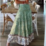 Photo d'une jeune femme debout devant une table rectangulaire avec des chaises portant une jupe bohême verte avec taille haute à smocks et large volant blanc en bas u sac blanc sur l'épaule