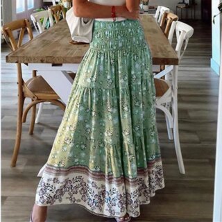 Photo d'une jeune femme debout devant une table rectangulaire avec des chaises portant une jupe bohême verte avec taille haute à smocks et large volant blanc en bas u sac blanc sur l'épaule