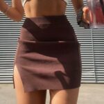 Photo d'une jeune femme debout devant un store baissé un verre à la main et portant une jupe courte marron fendue en tricot et un top blanc court