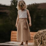 On voit une petite fille blonde d'environ 5 ans qui regarde sa jupe beige, de style minimaliste. Elle est dans un jardin, près d'un ballot de paille.