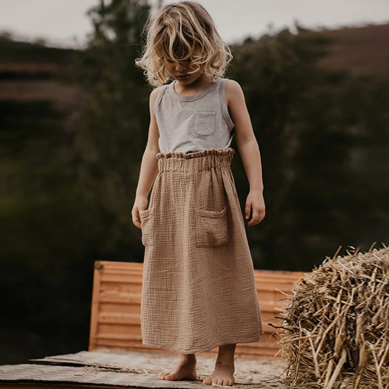 On voit une petite fille blonde d'environ 5 ans qui regarde sa jupe beige, de style minimaliste. Elle est dans un jardin, près d'un ballot de paille.