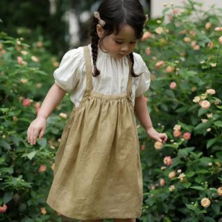 On voit une petite fille brune dans un jardin fleuri qui porte un chemisier et une jupe vert kaki.