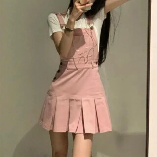 Photo d'une jeune femme aux cheveux longs debout le bras gauche levé et le droit plié portant une jupe salopette mini rose plissée