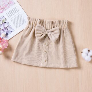 Sur fond rose, on voir une jupe trapèze beige pour petite fille. Elle est ornée d'un noeud de la même couleur et de faux boutons-pression sur le devant.