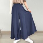 Photo d'une jeune femme debout portant une jupe culotte bleue large , des bakets blanches et un tee-shirt marron : sa main droite tient le côté de sa jupe