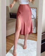Photo d'une femme debout dans un salon sur un tapis blanc et portant une jupe rose en satin à fente sur la jambe droite et un top blanc