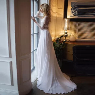 On voit une mariée qui porte un haut et une jupe de mariage blancs et qui regarde par la fenêtre. La pièce est cossue, elle se trouve dans un manoir ou un chaêteau.