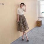 Femme adossée sur un mur extérieur beige qui porte une jupe dorée taille haute en Jacquard et un top blanc.