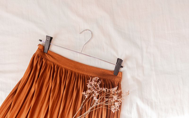 Haut de jupe plissée orange foncée, accroché à un cintre à pince, avec une fleur séché posé sur un tissu blanc.