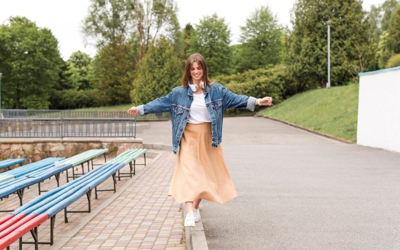 Femme habillée d'une jupe longue rose, orange avec un haut blanc et une veste en jean, elle porte des baskets blanches.