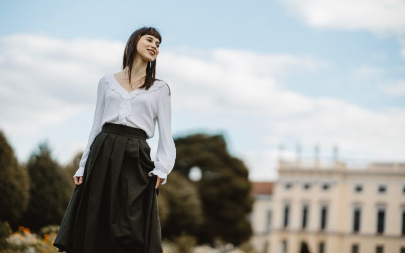 Femme vêtue d'une chemise blanche à manches longues et d'une jupe noire longue avec des plis plats. On voit un monument derrière elle.